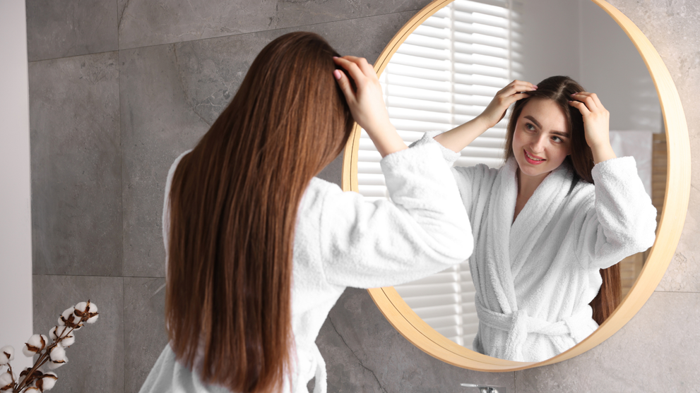 A woman examining her healthy, thick, long hair in a bathroom mirror, reflecting real results from consistent hair care.